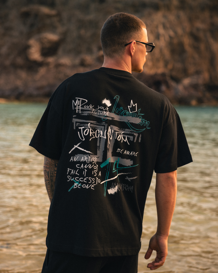 Man met zwarte T-shirt met kleurrijke tekst en grafische prints op de rug, staand bij het water met een rotsachtige achtergrond.