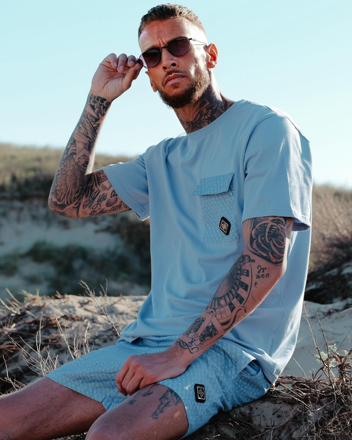 Man met tatoeages draagt lichtblauwe korte mouwen t-shirt en bijpassende korte broek, zittend in een natuurlijke buitenomgeving met zand en struiken, zonlicht zichtbaar.