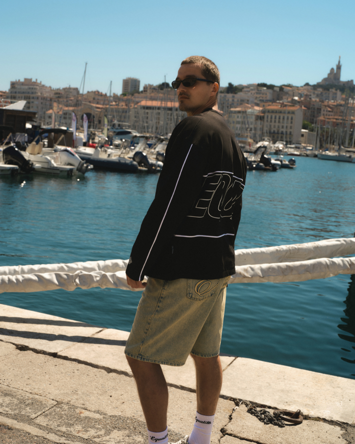 Man met een zwarte hoodie en beige korte broek staat bij een haven met boten en helder blauw water, omringd door stedelijke gebouwen op de achtergrond.