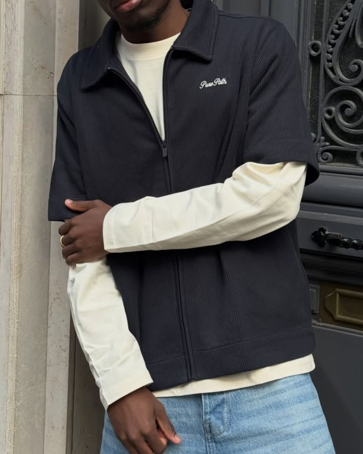 Man draagt een donkerblauwe korte mouw jas over een lichtgekleurde longsleeve en een lichte spijkerbroek, staand bij een klassieke deur met sierlijke details.