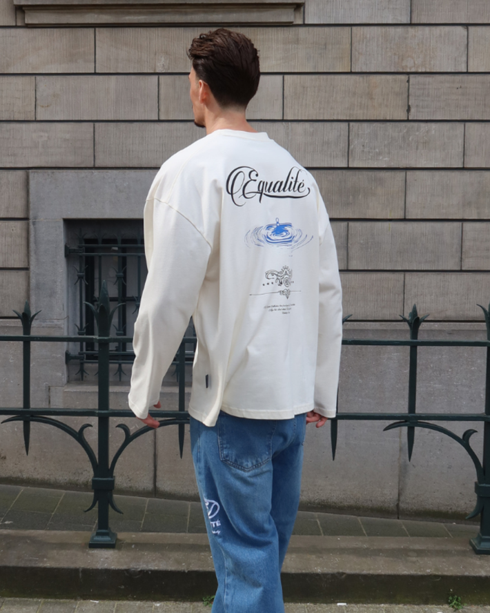 Man met donker haar draagt een witte longsleeve met tekst en afbeeldingen op de rug, gecombineerd met een blauwe losse spijkerbroek, staand voor een stenen muur met zwart hekwerk.