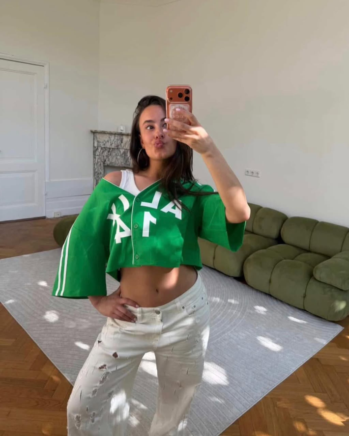 Vrouw die een groene cropped hoodie draagt met witte tekst en witte high waist broek met gaten, die een spiegelselfie maakt in een lichte kamer met een groene bank.