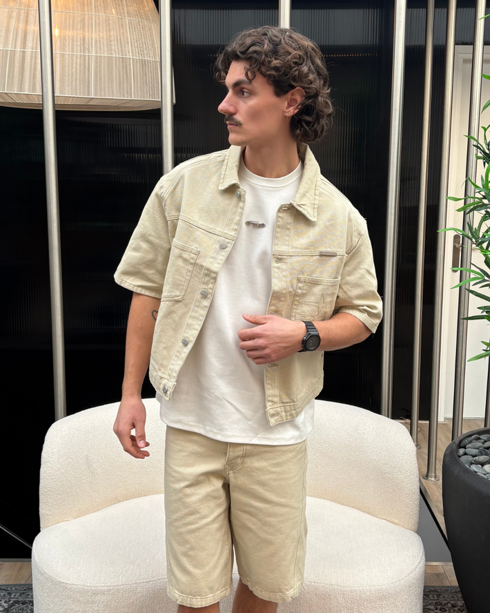 Man met krullend haar draagt een casual beige korte zomerjas en bijpassende korte broek met een wit T-shirt, staand in een moderne ruimte met bank en planten.