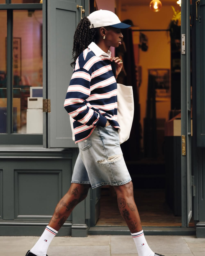 Vrouw loopt buiten in casual zomeroutfit met een gestreepte longsleeve, lichte denim korte broek, witte pet, sokken en sneakers, met een witte schoudertas.