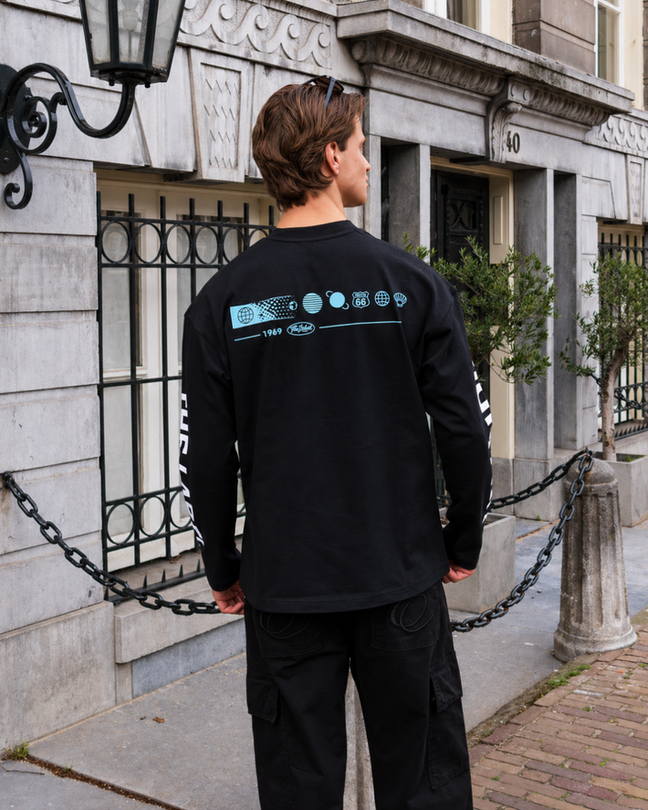 Man met bruin haar draagt een zwarte longsleeve met blauwe prints op de rug, staand op een straat voor een oud stenen gebouw met zwarte hekwerken en planten.