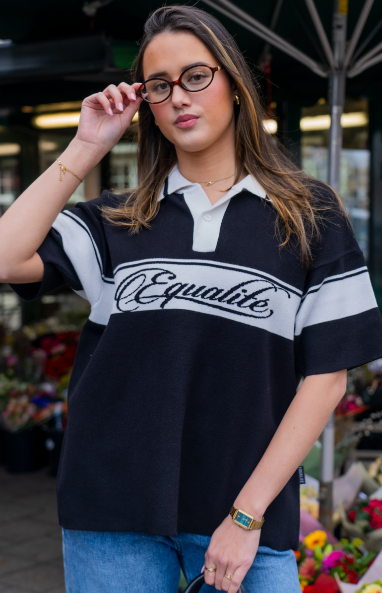 Vrouw draagt een zwarte polo met witte strepen en het woord Equalite in sierlijke letters, gecombineerd met een casual jeans en een bril.